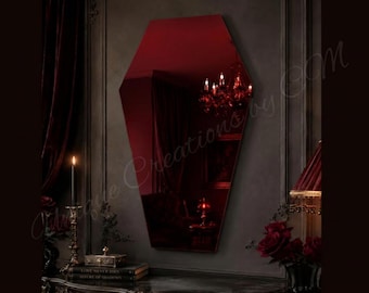 May include: A coffin-shaped wall mirror with a deep red tinted glass surface. The mirror features a geometric, six-sided silhouette with angled top and bottom edges, reflecting a dimly lit room with a chandelier and dark interior decor elements.