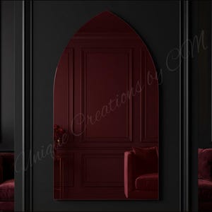 May include: A tall, arched mirror with a pointed top, featuring a deep burgundy tinted glass surface. The mirror reflects a room with dark walls and red furniture. The text Unique Creations by CM is overlaid across the center of the mirror.