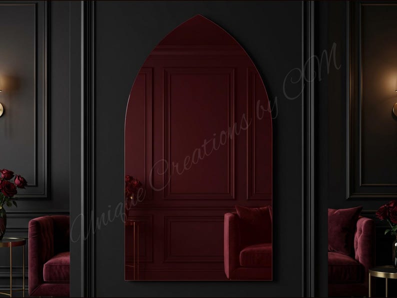 May include: A tall, arched mirror with a pointed top, featuring a deep burgundy tinted glass surface. The mirror reflects a room with dark walls and red furniture. The text Unique Creations by CM is overlaid across the center of the mirror.