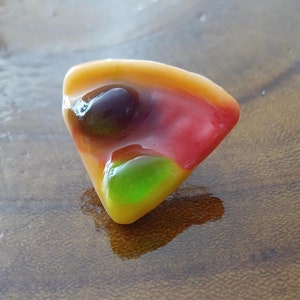 Puede incluir: Un caramelo gomoso con forma de porción de pizza con ingredientes rojos, amarillos y verdes.
