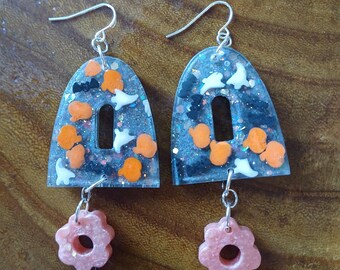May include: A pair of Halloween-themed earrings with a black and white glitter background. The earrings are shaped like arches with a small hole in the center. The arches are filled with orange and white Halloween-themed charms. The earrings are attached to a silver hook and dangle from a pink flower charm.