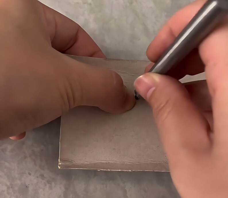 May include: A metallic gray stylus tool is being used to press a small, dark circular point onto the surface of a flat, square-shaped light gray material. The tool has a smooth, cylindrical body and a pointed tip for precise application.