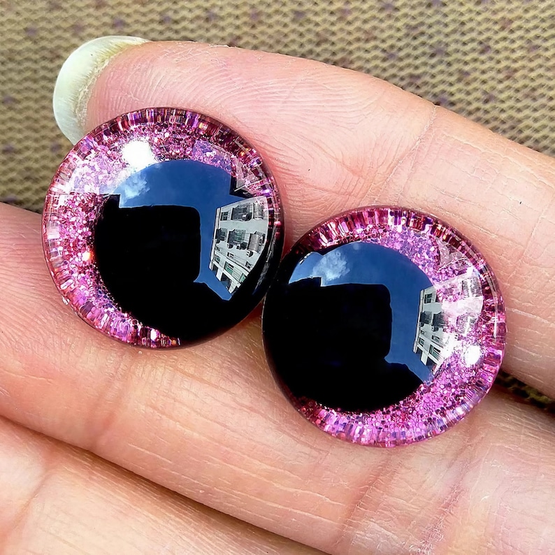 May include: Two black plastic eyes with a pink glitter rim. The eyes are round and have a reflective surface.