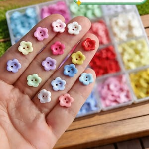 May include: A hand displays a variety of flower-shaped buttons in pastel colors, each approximately 10mm in diameter. The buttons are arranged against a backdrop of a clear plastic organizer filled with other colorful buttons. The buttons have two holes.