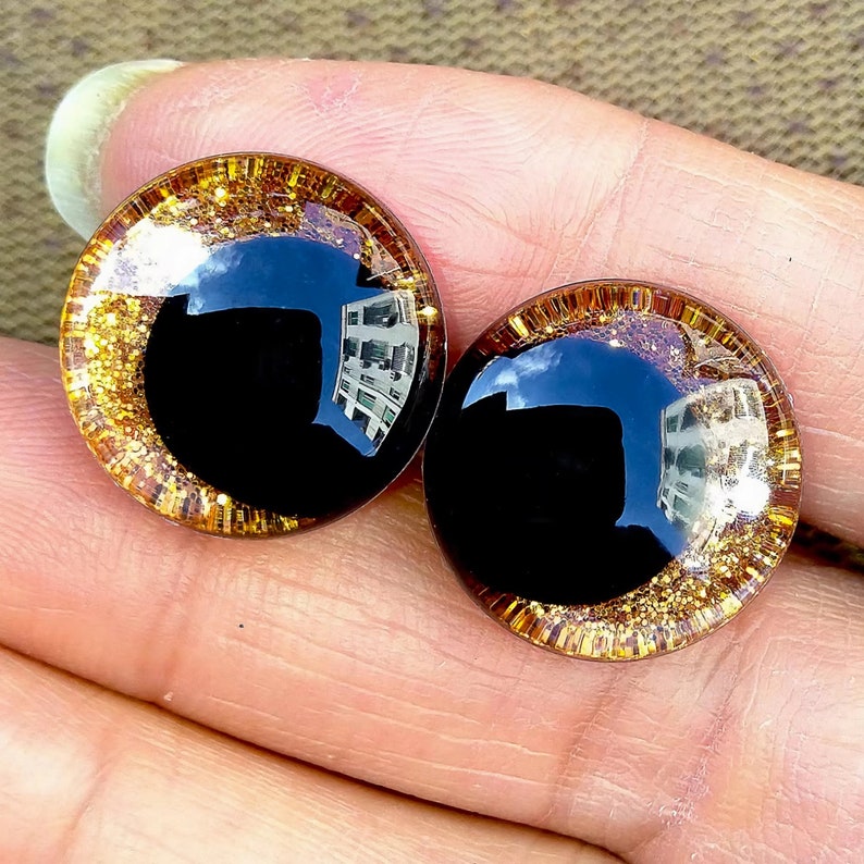 May include: Two large, round, black plastic eyes with a gold glitter rim. The eyes are on a hand.