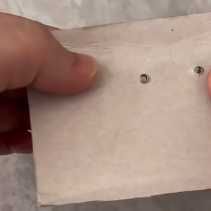 May include: Two small, circular, metallic stud earrings are attached to a rectangular piece of white cardboard. The earrings feature a simple, minimalist design with a polished finish, securely fastened through the paper backing for display.