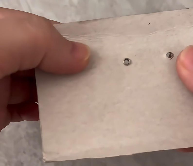 May include: Two small, circular, metallic stud earrings are attached to a rectangular piece of white cardboard. The earrings feature a simple, minimalist design with a polished finish, securely fastened through the paper backing for display.