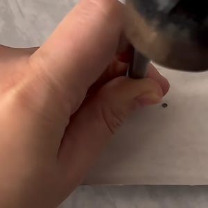May include: A metal hole punch tool with a dark cylindrical handle is being pressed down onto a flat, light-colored surface to create a small, circular perforation. The tool features a sturdy construction designed for manual pressure application.