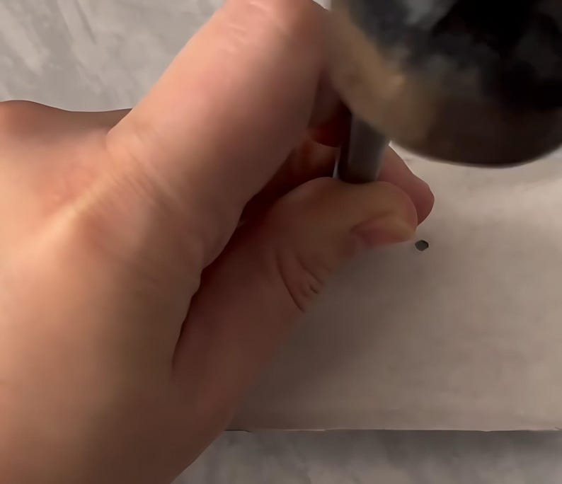May include: A metal hole punch tool with a dark cylindrical handle is being pressed down onto a flat, light-colored surface to create a small, circular perforation. The tool features a sturdy construction designed for manual pressure application.