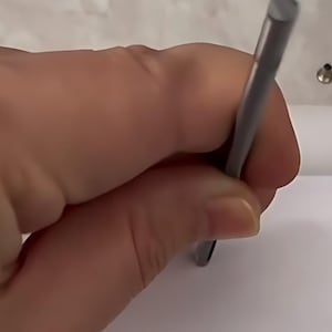 May include: A slender, cylindrical metal tool with a matte grey finish, held vertically against a plain white surface. The tool features a smooth, uniform body and a flat, circular tip, designed for precision tasks or marking on a flat material.