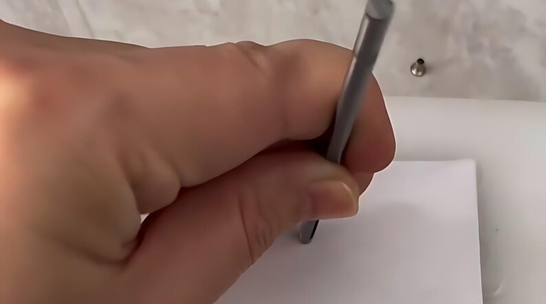 May include: A slender, cylindrical metal tool with a matte grey finish, held vertically against a plain white surface. The tool features a smooth, uniform body and a flat, circular tip, designed for precision tasks or marking on a flat material.