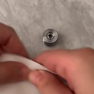 May include: A small, cylindrical silver metal component with a flat top surface featuring a centered circular indentation and a small dark hole in the middle. The object has a smooth, metallic finish and a compact, industrial design.