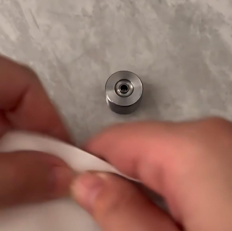 May include: A small, cylindrical silver metal component with a flat top surface featuring a centered circular indentation and a small dark hole in the middle. The object has a smooth, metallic finish and a compact, industrial design.
