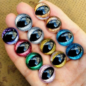 Könnte beinhalten: Sortiment von 10 bunten Glitzeraugen Bastelbedarf. Die Augen sind rund und haben eine schwarze Pupille. Die Farben umfassen Gold, Silber, Blau, Grün, Rosa und Rot.