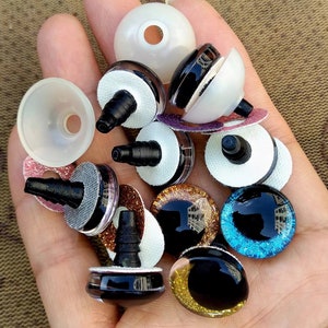 May include: Assortment of colorful plastic safety eyes for crafts. The eyes come in various sizes and colors, including black, brown, gold, and blue. Some eyes have glitter accents. The eyes are attached to a white backing with a black plastic post.
