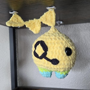 Puede incluir: Un juguete de peluche de ganchillo amarillo hecho a mano con un diseño negro y un ojo negro. El juguete tiene detalles en azul y verde en la parte inferior y cuelga de una estantería.