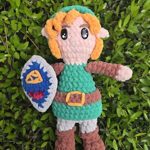 Puede incluir: Muñeca de ganchillo hecha a mano de un personaje con túnica verde, botas marrones y un escudo. La muñeca tiene pelo naranja, un gorro verde y un cinturón marrón. El escudo es azul, blanco y rojo.