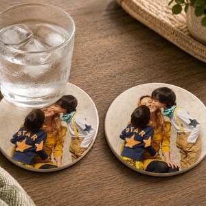 May include: Two round stone coasters featuring a printed photograph of a person being kissed on the cheek by a child wearing a navy blue shirt with a yellow star and another child wearing a white shirt with blue stars and yellow suspenders.