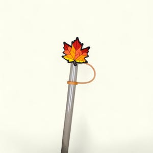 May include: A clear plastic straw with a fall-themed maple leaf topper. The leaf is in shades of red, orange, and yellow with a black outline. An orange silicone ring secures the straw. The straw is a reusable drinking accessory.