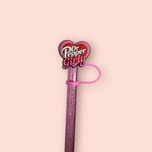 May include: A pink glitter straw with a pink heart-shaped charm that says "Dr. Pepper Girly". The straw has a pink silicone ring to prevent it from falling out of a cup. The background is a solid light pink.