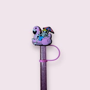 May include: A purple glitter straw with a skeleton riding a flamingo floatie. The skeleton is wearing a yellow hat and blue shorts, and is holding a red, white, and blue umbrella. A purple ring secures the straw.