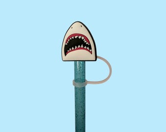 May include: A shark-themed straw topper with a white, black, and red design. The straw is blue with glitter and has a clear ring to hold the topper in place. The background is a solid light blue.