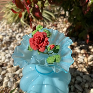 May include: A red beaded rose with green leaves sits in a light blue ruffled vase. The vase is on a bed of white pebbles.