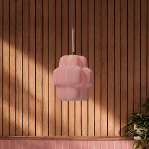Puede incluir: Una lámpara colgante de color rosa pálido con una forma geométrica redondeada y segmentada cuelga de un cable fino. La luminaria presenta una serie de estrías verticales y un acabado mate suave, suspendida sobre una mesa de madera y una zona de asientos de azulejos rosas.