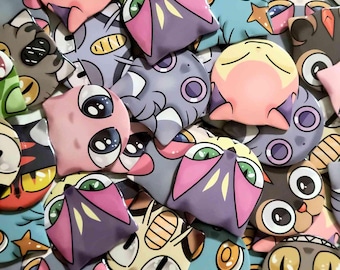 May include: A collection of colorful, cartoonish creature-shaped pins. The pins feature various designs with large eyes, in shades of pink, purple, blue, and yellow. Some have star and moon accents, and others have wing-like features.