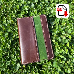 May include: A handcrafted leather wallet in shades of brown and green, with visible stitching. The wallet features a central green panel and a PDF download icon in the upper right corner. The item is set against a backdrop of green foliage.