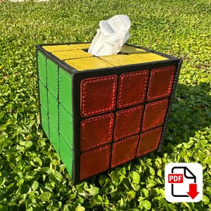 May include: A Rubik's Cube-themed tissue box with a black border, featuring yellow, red, and green squares. White tissues are visible at the top. The box is sitting on a bed of green foliage.