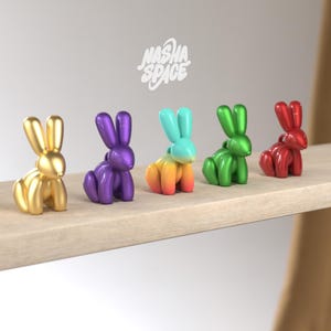 May include: Five balloon animal rabbit figurines in different colours: gold, purple, orange and blue gradient, green, and red. The figurines are arranged on a wooden shelf. The text "nasha space" is written in white on a white background above the figurines.