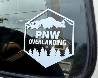 May include: White vinyl sticker with a hexagonal shape. The sticker features a mountain range silhouette, the text "PNW OVERLANDING", and a row of evergreen trees at the bottom.