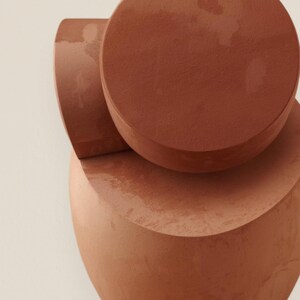 Pode incluir: Escultura abstrata em terracota composta por formas geométricas empilhadas. Uma base arredondada suporta um elemento curvo semicircular, com uma forma circular no topo. Os tons quentes e terrosos e as superfícies lisas criam uma estética moderna e minimalista.