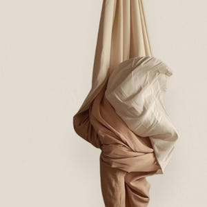 May include: Folded fabric in beige and brown tones hangs against a neutral backdrop. The textiles are gathered and knotted, creating a soft, textured effect. The image showcases the interplay of light and shadow on the fabric, highlighting its texture.