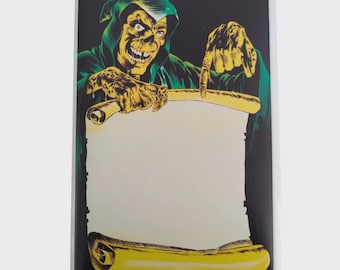 Op de afbeelding: Een rechthoekige sticker met een skeletachtige figuur in een groene capuchon, die naar een blanco rol wijst. De figuur heeft gele huid en een sinistere grijns. De rol is geel met een wit midden, tegen een zwarte achtergrond.