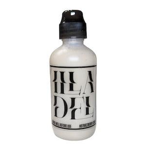 Puede incluir: Una botella blanca con tapa negra y una etiqueta con la palabra "HILADEL" en letras negras en negrita y estilizadas. La etiqueta también incluye el texto "SHAKE WELL BEFORE USE" y "HOTRAZZBERRY DRINK". La botella contiene un líquido blanco.