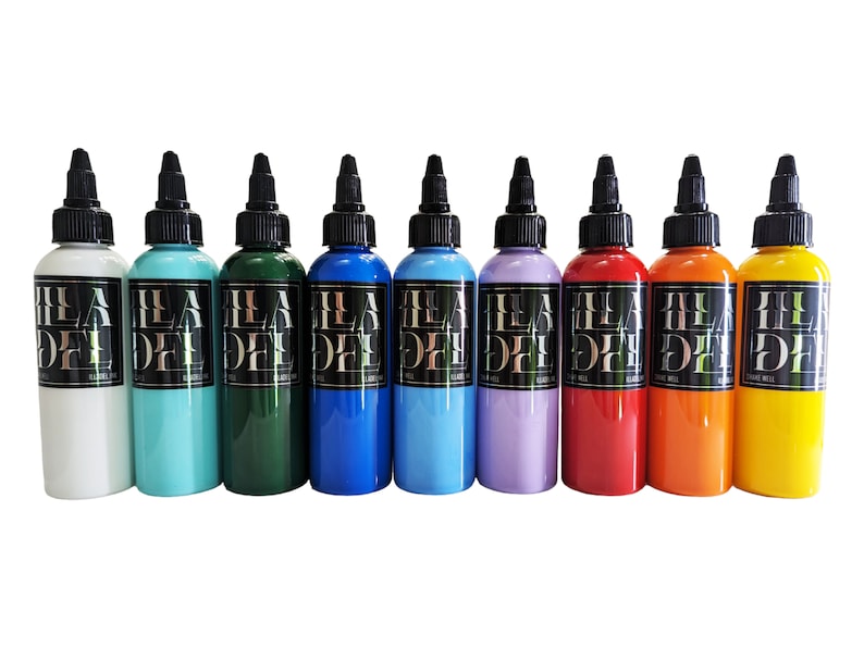 May include: Ten bottles of tattoo ink in various colours, including white, turquoise, green, blue, light blue, lavender, red, orange, and yellow. Each bottle has a black label with the text "ILA DYE" in white.