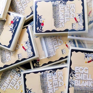 May include: Stacks of cream-colored Egg Shell Blank stickers with a black border and cartoon graphics. The stickers feature a character holding a lit stick of dynamite. Text includes "Egg Shell Blanks" and "Super Permanent".