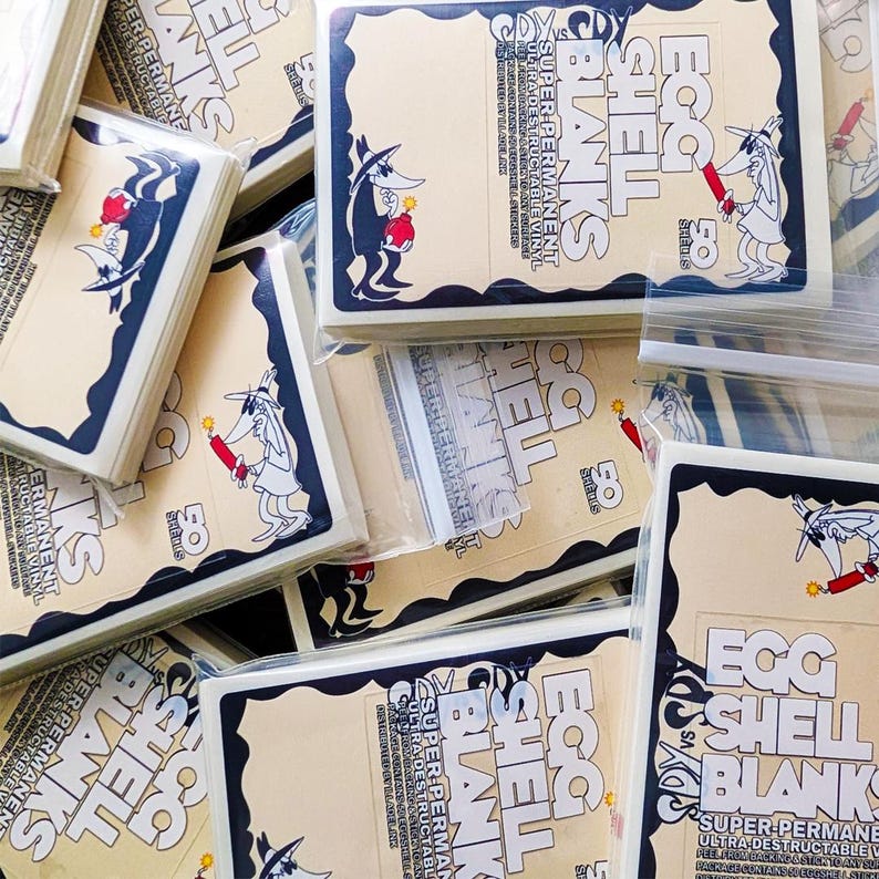 May include: Stacks of cream-colored Egg Shell Blank stickers with a black border and cartoon graphics. The stickers feature a character holding a lit stick of dynamite. Text includes "Egg Shell Blanks" and "Super Permanent".