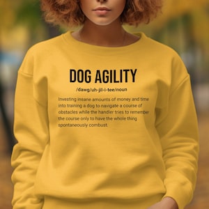Op de afbeelding: Geel sweatshirt met de tekst "DOG AGILITY" en een definitie van het woord. De definitie luidt: /dawg/uh-jil-i-tee/zelfstandig naamwoord Een krankzinnig bedrag aan geld en tijd investeren in het trainen van een hond om een parcours te navigeren, terwijl de handler probeert om het parcours te onthouden, alleen om te zien hoe het geheel spontaan in brand vliegt.