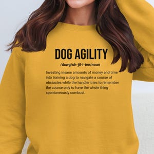 May include: Mustard yellow sweatshirt with the text "DOG AGILITY" and a definition. The definition describes the process of training a dog to navigate obstacles. The sweatshirt is a crew neck style.