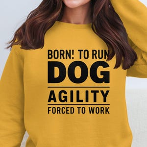 May include: Mustard yellow sweatshirt with the text "BORN! TO RUN DOG AGILITY FORCED TO WORK" in black. The sweatshirt is a solid color and has a crew neck.