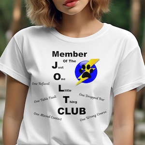 May include: White t-shirt with black text that reads "Member of the JOLT CLUB". The text is broken down into the letters J, O, L, T, and CLUB, with a description of what each letter stands for below it. The J stands for "Just", the O stands for "One", the L stands for "Little", the T stands for "Thing", and the CLUB is written in all caps. There is a blue circle with a yellow lightning bolt and a paw print in the centre of the shirt.