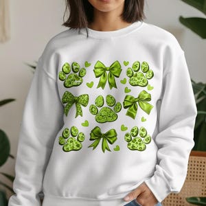 Op de afbeelding: Witte sweatshirt met een patroon van groene pootafdrukken, strikken en hartjes. De pootafdrukken hebben een gespikkeld ontwerp en de strikken zijn effen groen. Kleine groene hartjes zijn verspreid.