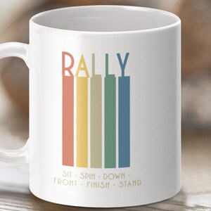 May include: White ceramic mug with a colorful graphic that says "RALLY" with the words "SIT - SPIN - DOWN - FRONT - FINISH - STAND" underneath.
