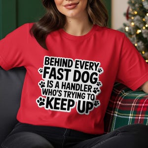 May include: A red t-shirt with the text "BEHIND EVERY FAST DOG IS A HANDLER WHO'S TRYING TO KEEP UP" in a white, paw-print-decorated design. The shirt is being worn by a person sitting on a couch.