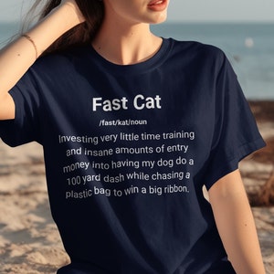 May include: A navy blue t-shirt with white text that reads "Fast Cat /fast/kat/noun Investing very little time training and insane amounts of entry money into having my dog do a 100 yard dash while chasing a plastic bag to win a big ribbon."