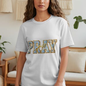 Könnte beinhalten: Weißes T-Shirt mit dem Text "Just Pray" in einem floralen und goldenen Design. Der Text ist in einer stilisierten Schriftart mit blauen und gelben Blumenmustern gehalten. Das Shirt hat einen Rundhalsausschnitt und kurze Ärmel.