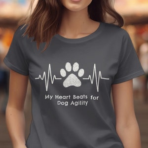 Puede incluir: Una camiseta gris con una huella de pata blanca y un gráfico de latido del corazón. El texto "My Heart Beats for Dog Agility" está impreso debajo del gráfico.